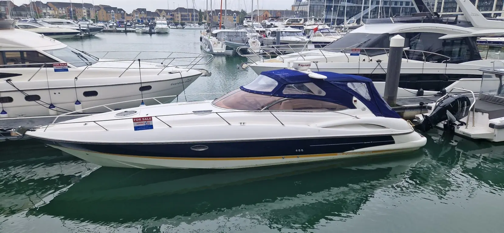 A white motorboat with a blue canopy is docked at a marina, displaying a "For Sale" sign on the side.